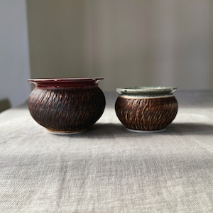 May include: Two brown ceramic pots with a textured surface. The larger pot has a dark red rim and the smaller pot has a light green rim.