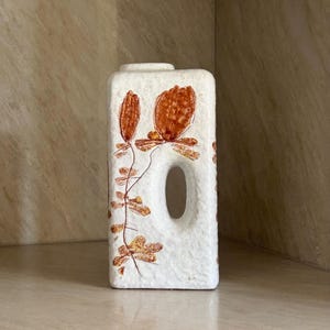May include: A rectangular white ceramic vase with a textured, matte finish. The vase features a central vertical oval cutout and is decorated with a brown botanical illustration of stems and leaves running along the left side and top edge.