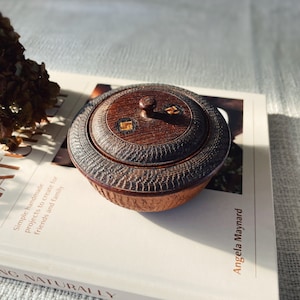 May include: A small, round, wooden box with a lid. The box is dark brown with a textured surface. The lid has a small knob on top and is decorated with a geometric pattern in gold.