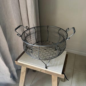 May include: A black metal wire basket with two handles sits on a wooden stool. The basket has a round shape and a wire mesh design.