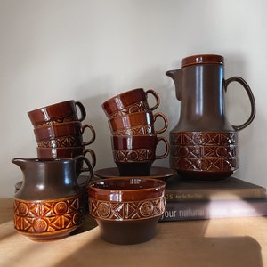 May include: A brown ceramic coffee set with a geometric design, including a coffee pot, creamer, sugar bowl, cups, and saucers. The cups are stacked. The set is displayed on a wooden surface. The coffee pot and sugar bowl have a similar design.