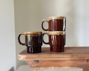 May include: Three stacked brown ceramic mugs with a textured design. The mugs have a dark brown base and a lighter brown rim. The mugs are on a wooden surface.