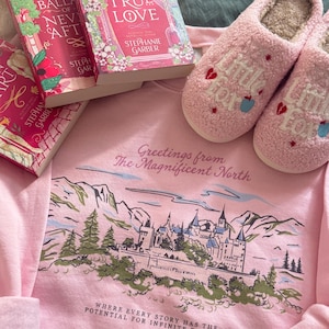 Puede incluir: Una sudadera rosa con un gráfico de castillo y el texto "Greetings from The Magnificent North" y "Where every story has the potential for infinite endings." También, dos zapatillas rosas y tres libros son visibles.