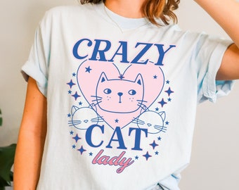 May include: A light blue t-shirt with a pink heart and a cartoon cat inside. The text "CRAZY CAT lady" is printed on the shirt in blue.