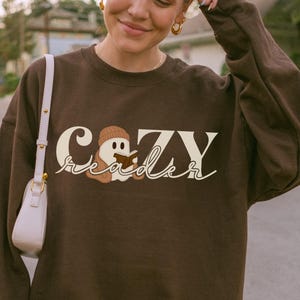 May include: A brown sweatshirt featuring the text "Cozy Reader" in a playful font. The design incorporates a cartoon ghost wearing a beanie. The sweatshirt is a plain brown colour.