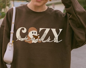 May include: A brown sweatshirt with the words "Cozy Reader" in a whimsical font. The design includes a cartoon ghost wearing a beanie. The sweatshirt is a solid brown color.