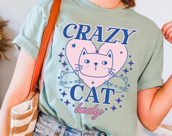 May include: A light blue t-shirt with a pink heart and a cartoon cat inside. The text "CRAZY CAT lady" is printed on the shirt in blue.
