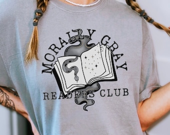 May include: A grey t-shirt featuring a graphic design of a book with a snake and stars. The text "Morally Gray Readers Club" is printed on the shirt.