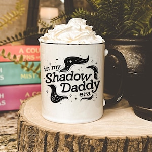 Puede incluir: Taza de cerámica blanca con borde y asa negros. La taza tiene un gráfico negro que dice "in my Shadow Daddy era" con estrellas y un diseño de nube negra estilizado.
