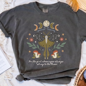 May include: A dark gray t-shirt with a graphic print of a moth, moon phases, and mushrooms. The text "For the girl whose eyes always stray to the Moon" is printed below the graphic.