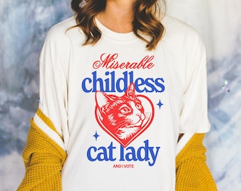Op de afbeelding: Crèmekleurig shirt met lange mouwen met de tekst "Miserable childless cat lady" in rood en blauw, met een kattenillustratie in een hart. Het shirt bevat ook de tekst "AND I VOTE".