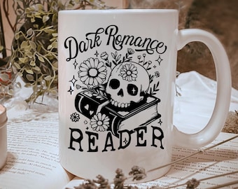 Può includere: Tazza in ceramica bianca con la scritta "Dark Romance Reader" in nero e un disegno di teschio con fiori. La tazza è appoggiata su un libro, con fiori secchi e una candela sullo sfondo.