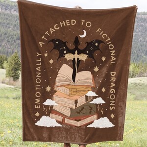 May include: A brown fleece blanket with the text "Emotionally Attached to Fictional Dragons" and a silhouette of a dragon flying over a stack of books.