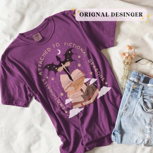 May include: Purple t-shirt with a graphic of a dragon flying over a stack of books. The text on the shirt reads "Emotionally attached to fictional dragons".