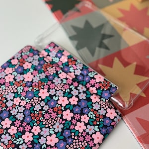 May include: A clear phone case and a floral patterned case with pink, purple, and blue flowers on a black background. The cases are on a white surface with a colorful starburst pattern in the background.