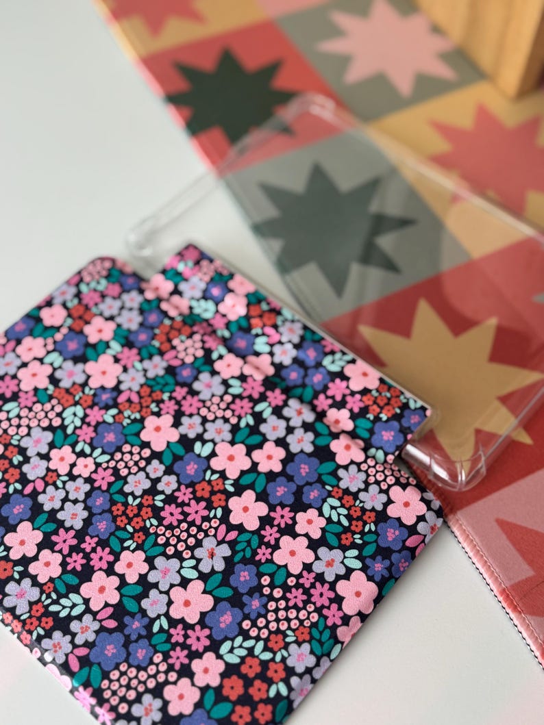 May include: A clear phone case and a floral patterned case with pink, purple, and blue flowers on a black background. The cases are on a white surface with a colorful starburst pattern in the background.
