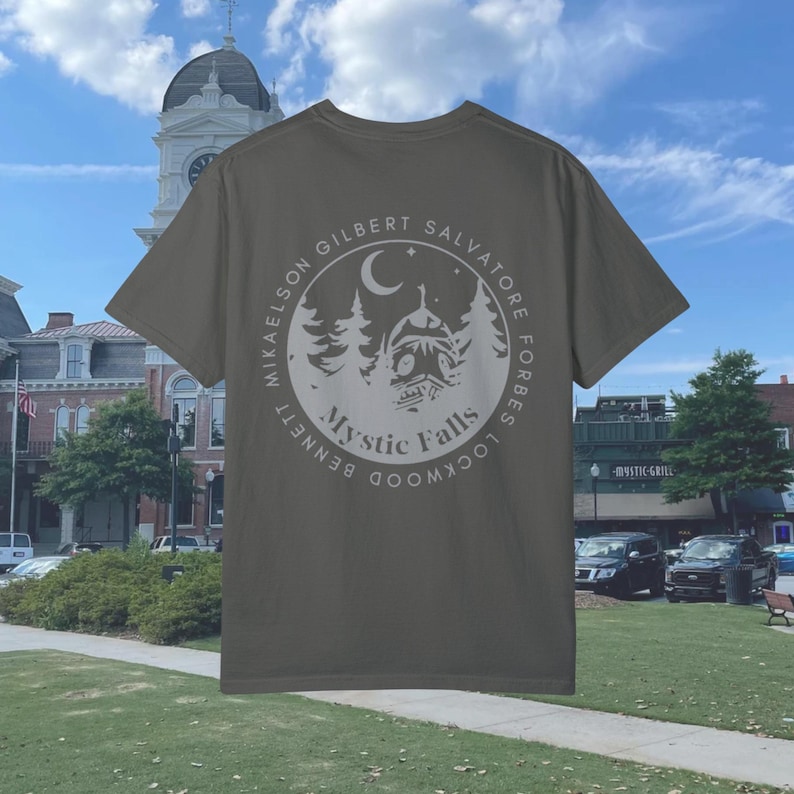Puede incluir: Una camiseta verde oscuro con un gr&aacute;fico circular blanco en la espalda. El gr&aacute;fico presenta una luna creciente, estrellas, &aacute;rboles, una casa y el texto "Mystic Falls" en un c&iacute;rculo con los nombres "Lockwood, Forbes, Salvatore, Gilbert, Mikaelson, Bennett" alrededor del exterior.