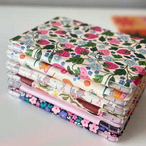 May include: A stack of fabric-covered phone cases. The top case features a cream background with pink strawberries, green leaves, and blue flowers. Other cases display various floral and patterned designs, including a pink satin edge on one.