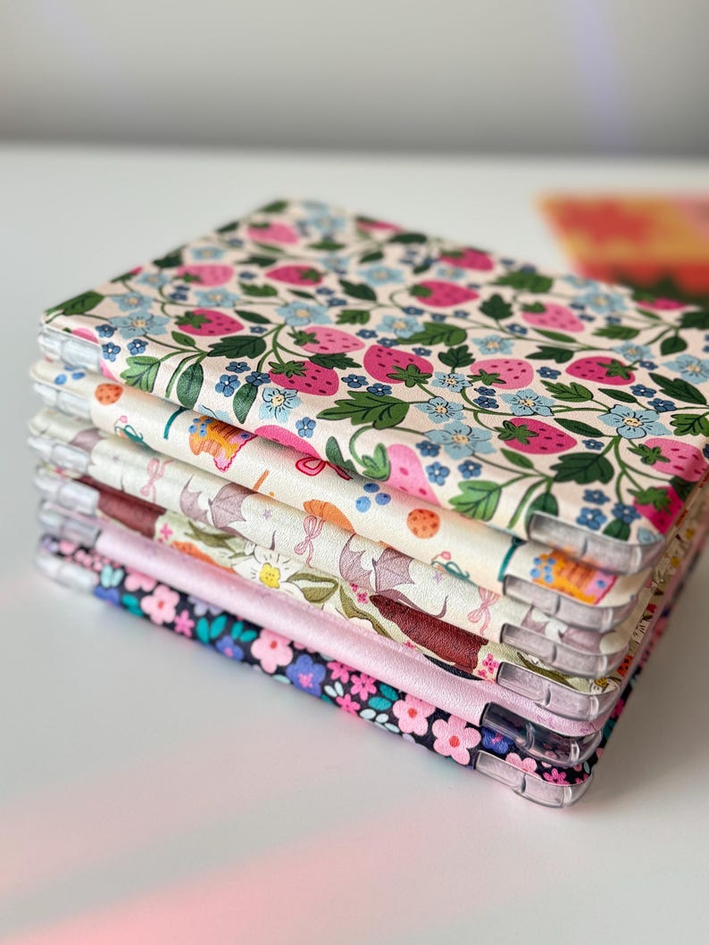 May include: A stack of fabric-covered phone cases. The top case features a cream background with pink strawberries, green leaves, and blue flowers. Other cases display various floral and patterned designs, including a pink satin edge on one.