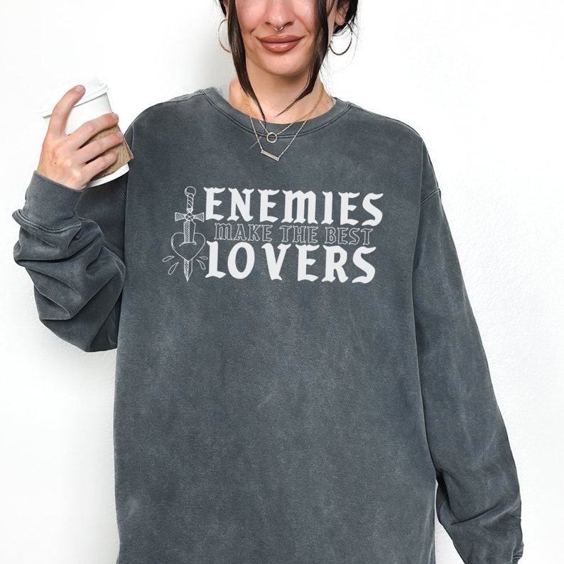 May include: A dark grey sweatshirt featuring the white text "ENEMIES MAKE THE BEST LOVERS". The design includes a sword and heart. The sweatshirt has a faded, vintage look.