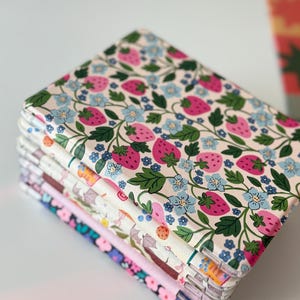 May include: A stack of fabric-covered notebooks with a strawberry and floral pattern. The notebooks feature a cream background with pink strawberries, blue flowers, and green leaves. Other notebooks in the stack have different floral patterns.