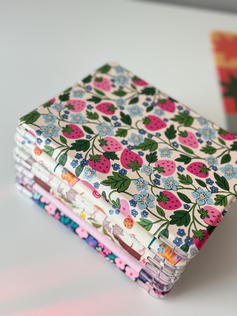 May include: A stack of fabric-covered notebooks with a strawberry and floral pattern. The notebooks feature a cream background with pink strawberries, blue flowers, and green leaves. Other notebooks in the stack have different floral patterns.