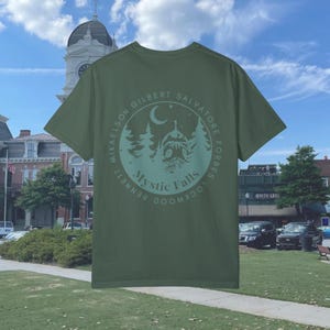 Puede incluir: Una camiseta verde con un gr&aacute;fico circular en la espalda. El gr&aacute;fico presenta una luna creciente, estrellas, &aacute;rboles y una casa con el texto "Mystic Falls" y los nombres de los personajes principales de la serie de televisi&oacute;n "The Vampire Diaries".
