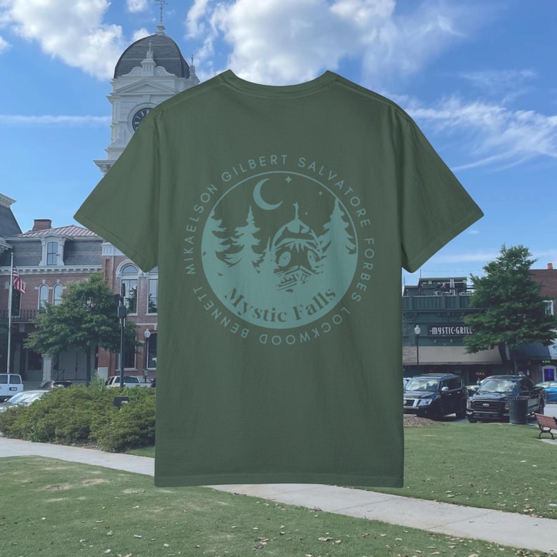 Puede incluir: Una camiseta verde con un gr&aacute;fico circular en la espalda. El gr&aacute;fico presenta una luna creciente, estrellas, &aacute;rboles y una casa con el texto "Mystic Falls" y los nombres de los personajes principales de la serie de televisi&oacute;n "The Vampire Diaries".