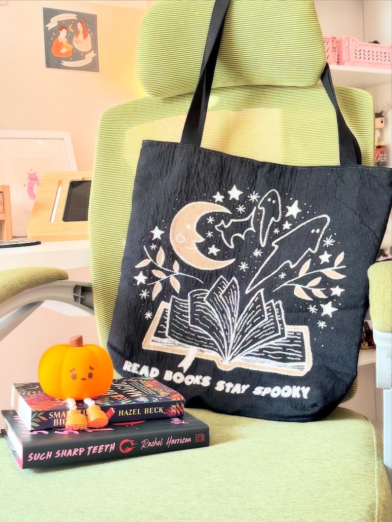 May include: Black tote bag with a spooky book design. The design features a white open book with a moon, stars, and ghosts. The text 'READ BOOKS STAY SPOOKY' is printed below. The bag is on a green chair with a pumpkin and books.