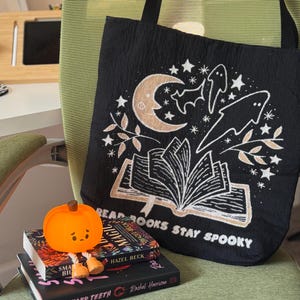 Puede incluir: Bolso tote negro con un dise&ntilde;o espeluznante que presenta una luna creciente, estrellas, fantasmas y un libro abierto. El texto "READ BOOKS STAY SPOOKY" est&aacute; impreso en la bolsa. Una peque&ntilde;a figura de calabaza naranja se encuentra encima de una pila de libros.