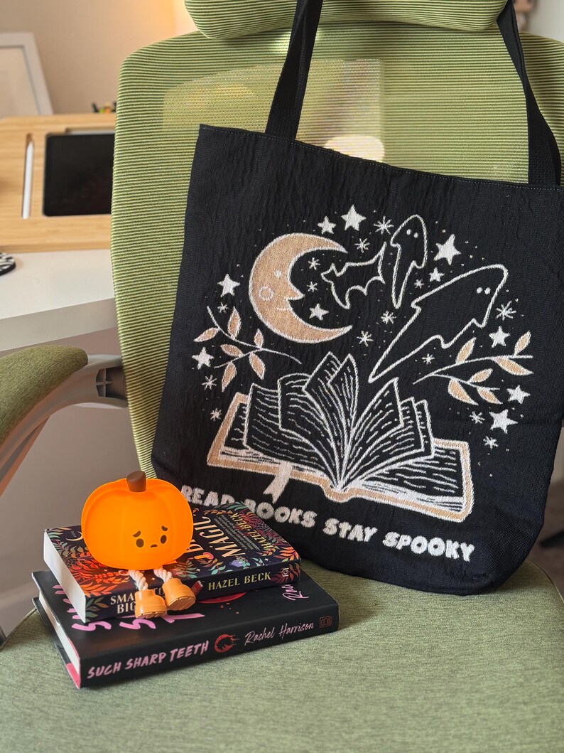 Puede incluir: Bolso tote negro con un dise&ntilde;o espeluznante que presenta una luna creciente, estrellas, fantasmas y un libro abierto. El texto "READ BOOKS STAY SPOOKY" est&aacute; impreso en la bolsa. Una peque&ntilde;a figura de calabaza naranja se encuentra encima de una pila de libros.