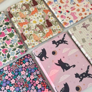 May include: A collection of patterned cases, featuring various designs. One case displays a pink background with black cat silhouettes, moons, and stars. Other cases showcase floral, strawberry, and coffee-themed patterns. The cases are likely for electronic devices.