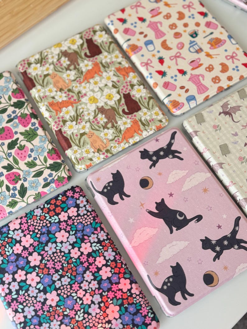 May include: A collection of patterned cases, featuring various designs. One case displays a pink background with black cat silhouettes, moons, and stars. Other cases showcase floral, strawberry, and coffee-themed patterns. The cases are likely for electronic devices.