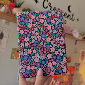 May include: A rectangular case with a floral pattern. The case features a dark background covered in small pink, blue, and orange flowers with green leaves. The case is held by a person wearing a blue sweater.