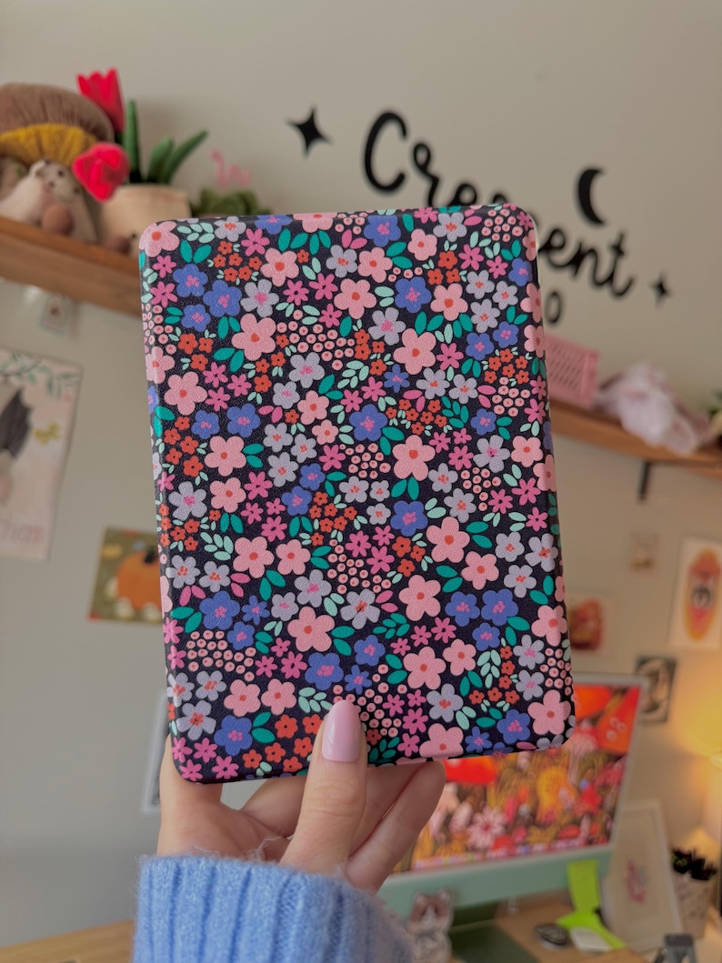 May include: A rectangular case with a floral pattern. The case features a dark background covered in small pink, blue, and orange flowers with green leaves. The case is held by a person wearing a blue sweater.
