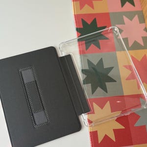 May include: A black tablet case with a gray elastic hand strap is open next to a clear protective case. A colorful patterned mat with star shapes in red, green, and pink is in the background.