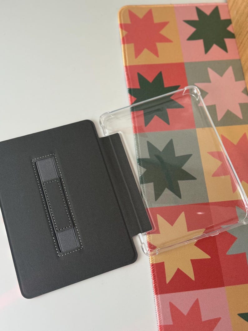 May include: A black tablet case with a gray elastic hand strap is open next to a clear protective case. A colorful patterned mat with star shapes in red, green, and pink is in the background.