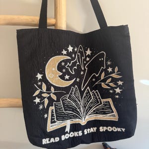 Puede incluir: Bolso tote negro con un dise&ntilde;o espeluznante que presenta una luna creciente, estrellas, fantasmas y un libro abierto. El texto "READ BOOKS STAY SPOOKY" est&aacute; impreso en dorado. El bolso tiene asas negras.