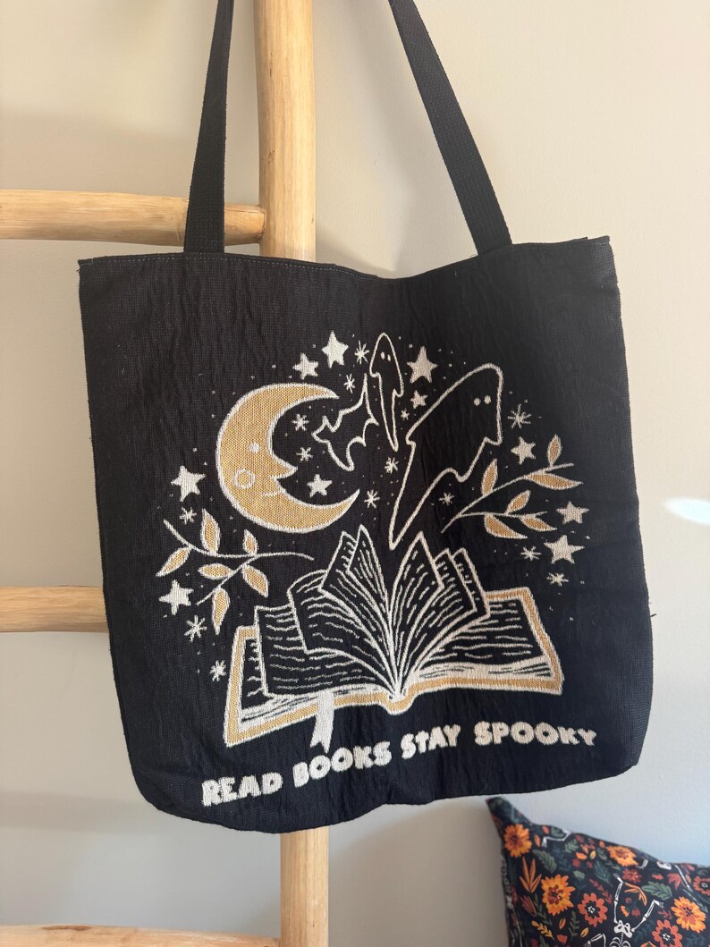 Puede incluir: Bolso tote negro con un dise&ntilde;o espeluznante que presenta una luna creciente, estrellas, fantasmas y un libro abierto. El texto "READ BOOKS STAY SPOOKY" est&aacute; impreso en dorado. El bolso tiene asas negras.
