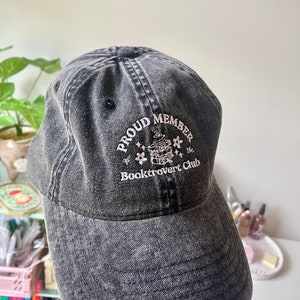 May include: A black denim baseball cap with the text "Proud Member of the Booktrovert Club" embroidered on the front. The text is in white and the embroidery includes a small illustration of a book with a flower on top.