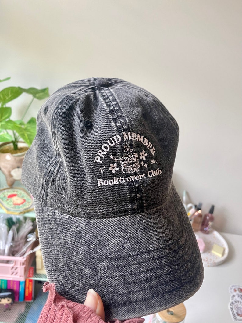 May include: A black denim baseball cap with the text "Proud Member of the Booktrovert Club" embroidered on the front. The text is in white and the embroidery includes a small illustration of a book with a flower on top.
