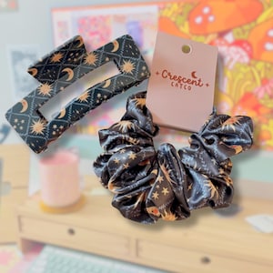 May include: A black rectangular hair claw clip and a matching black scrunchie, both featuring a celestial pattern of gold suns, moons, and stars. The scrunchie is attached to a light pink display card with the text Crescent CATCO printed on it.