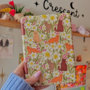 May include: A patterned book cover featuring a repeating design of cats and flowers. The cats are in shades of orange, brown, and burgundy, set against a backdrop of white flowers and green leaves. The text "Crescent" is visible on the wall.