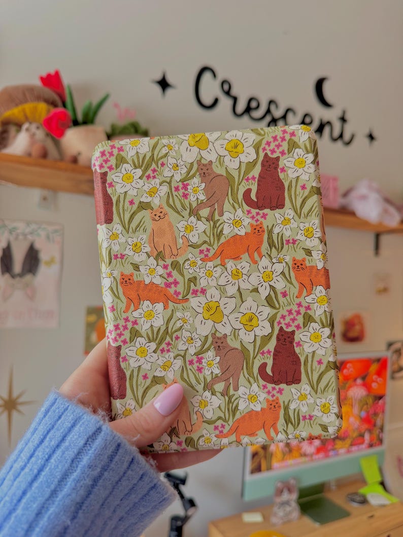 May include: A patterned book cover featuring a repeating design of cats and flowers. The cats are in shades of orange, brown, and burgundy, set against a backdrop of white flowers and green leaves. The text "Crescent" is visible on the wall.