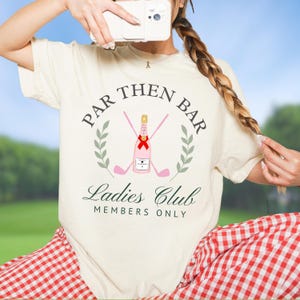 Op de afbeelding: Crèmekleurig T-shirt met een grafisch ontwerp. Het ontwerp bevat de tekst "Par Then Bar Ladies Club Members Only", een roze champagnefles, gekruiste golfclubs en een lauwerkrans. Het shirt heeft een relaxte pasvorm.