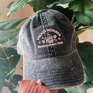 May include: A black denim baseball cap with a pink embroidered design that says "Proud Member of the Booktrovert Club".