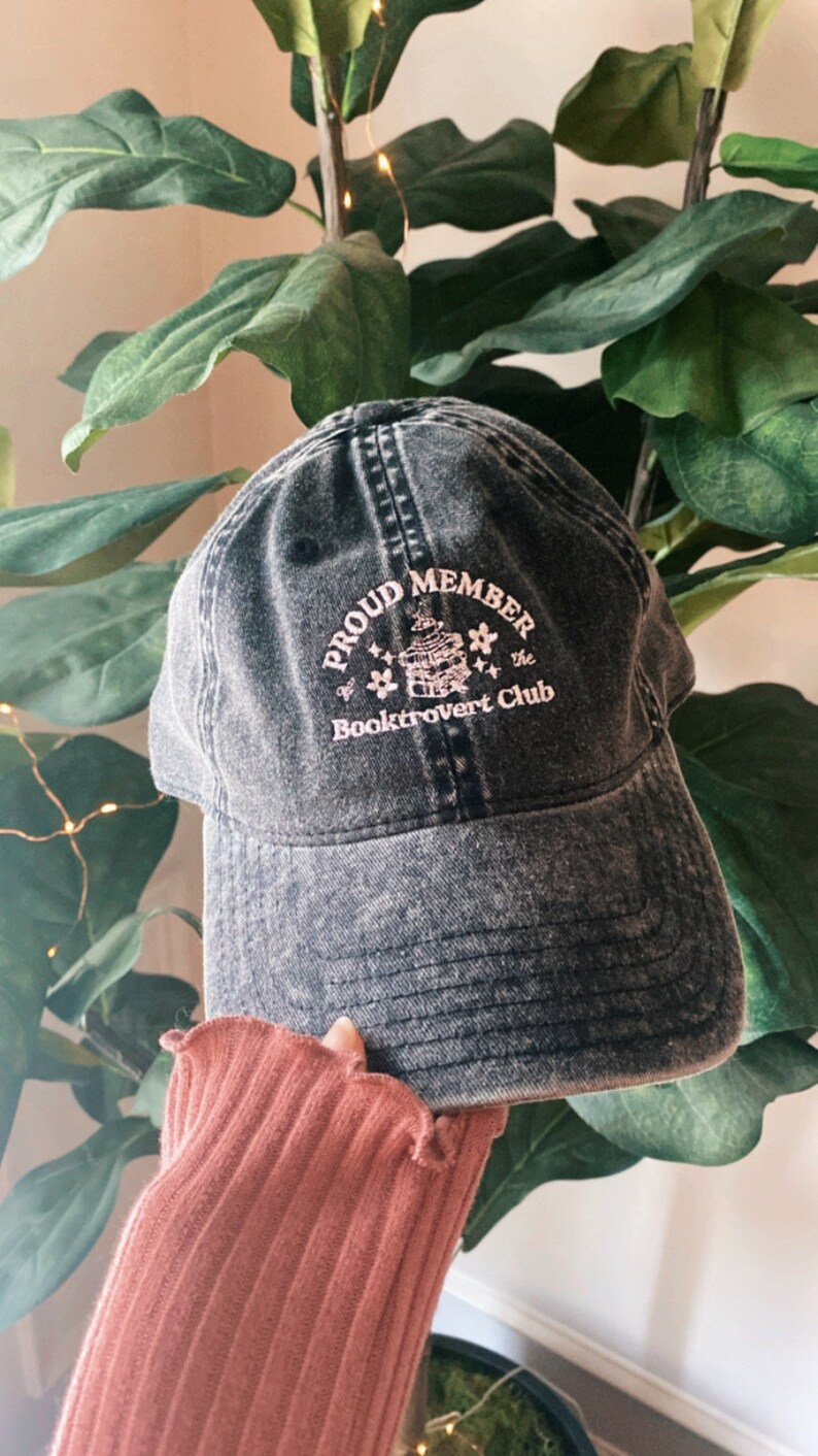 May include: A black denim baseball cap with a pink embroidered design that says "Proud Member of the Booktrovert Club".