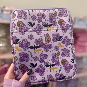 May include: A lavender tablet case with a repeating pattern of purple flowers, bats, dragons, and orange slices. The case has a zipper closure and a solid lavender border.