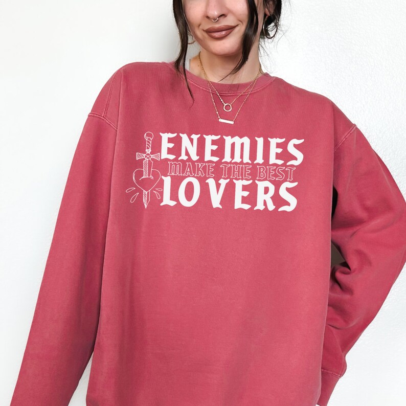 May include: A red-toned sweatshirt featuring the white text "ENEMIES MAKE THE BEST LOVERS" and a heart-and-dagger graphic. The crew neck sweatshirt has long sleeves.