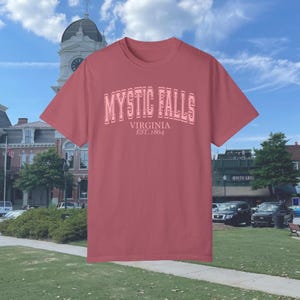 Puede incluir: Una camiseta roja con un gr&aacute;fico rosa que dice "Mystic Falls, Virginia Est. 1864".